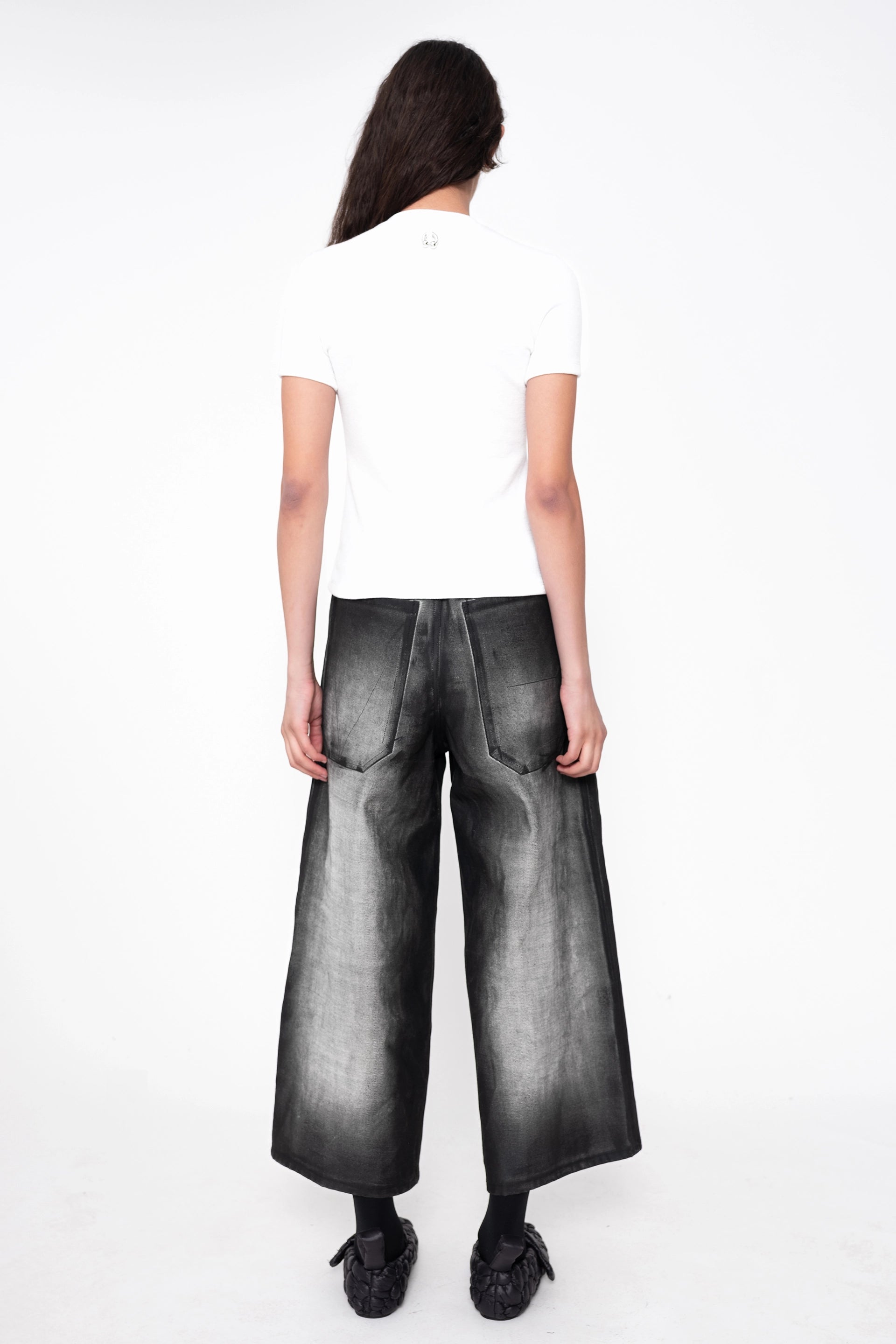 female-wearing-white-tee-and-cropped-grey-denim-pants-with-black-spray-paint-on-white-background-full-body-back