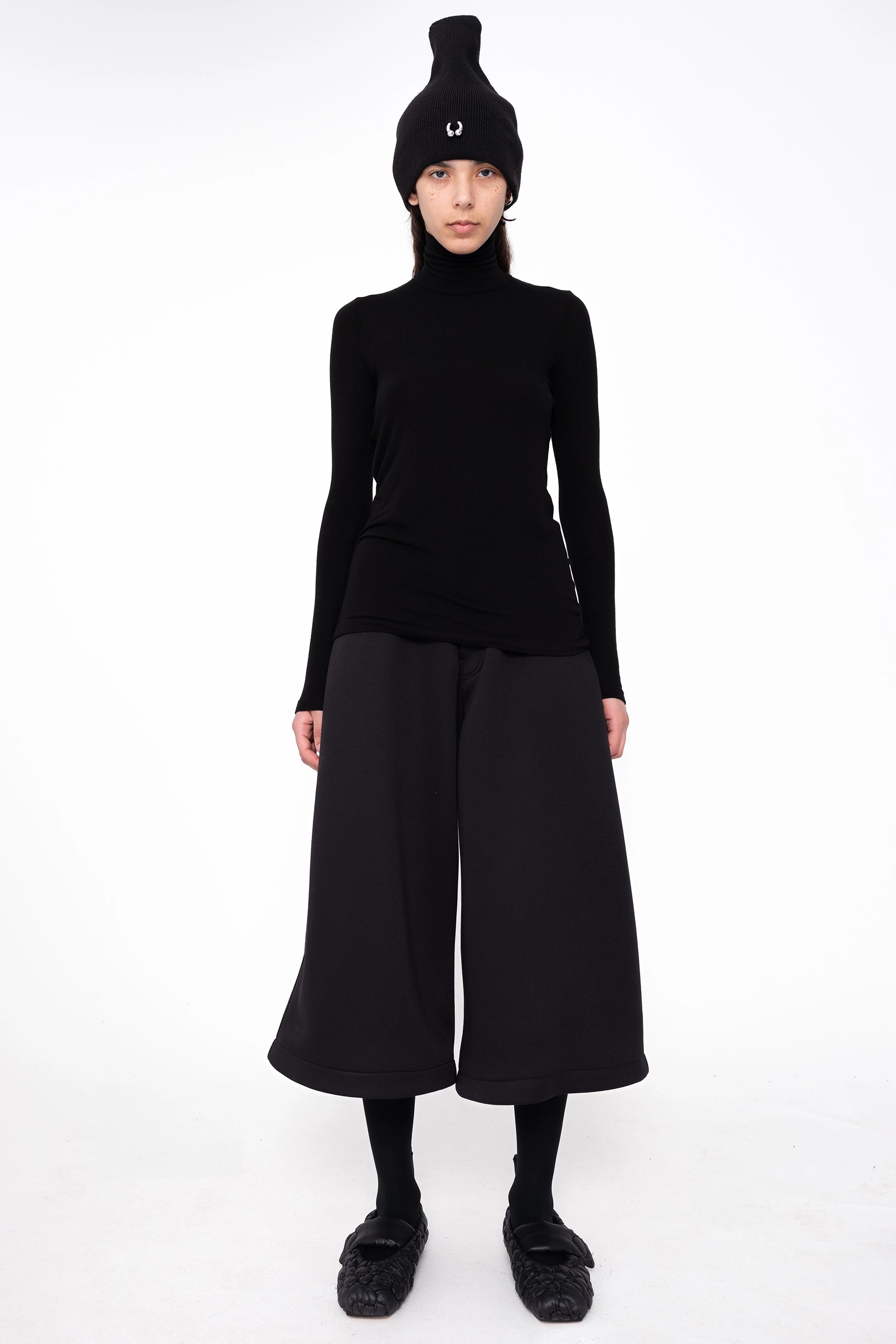 female-wearing-black-turtleneck-and-pierced-beanie-withwide-leg-culottes-on-white-background-full-body-front