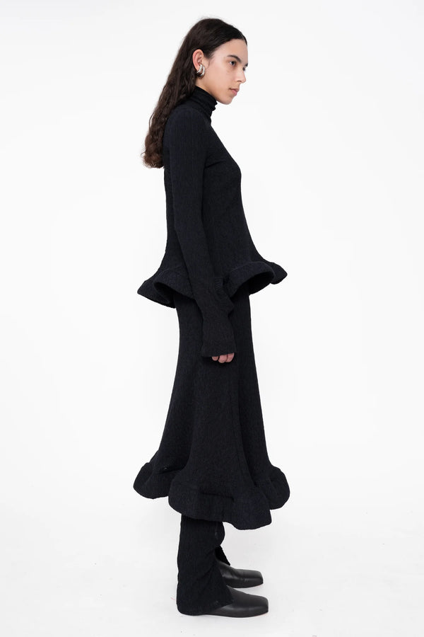 female-wearing-black-turtleneck-with-wavy-hem-and-matching-midi-skirt-on-white-background-full-body-side