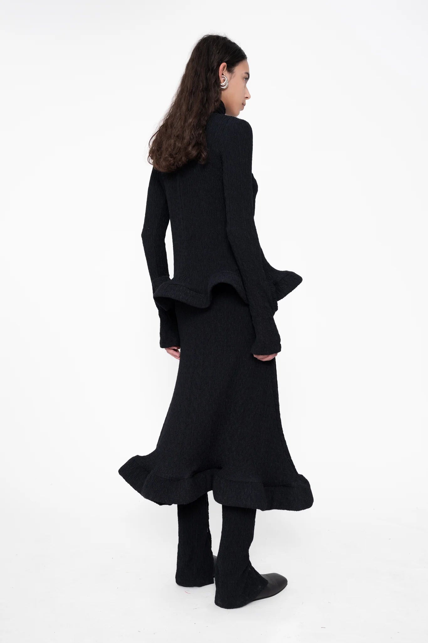 female-wearing-black-turtleneck-with-wavy-hem-and-matching-midi-skirt-on-white-background-full-body-back-side