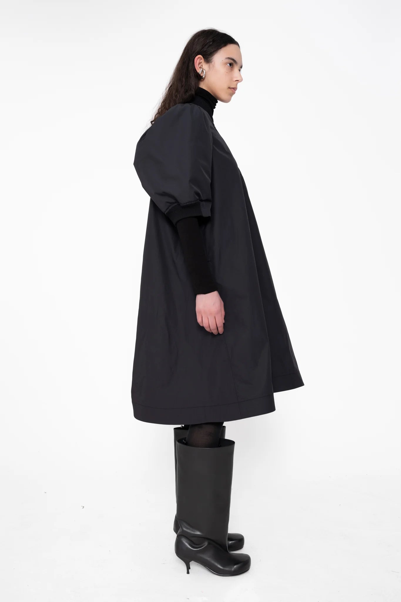 female-wearing-black-bomber-dress-with-zip-and-puffed-cropped-sleeves-and-double-heel-boots-on-white-background-full-body-side