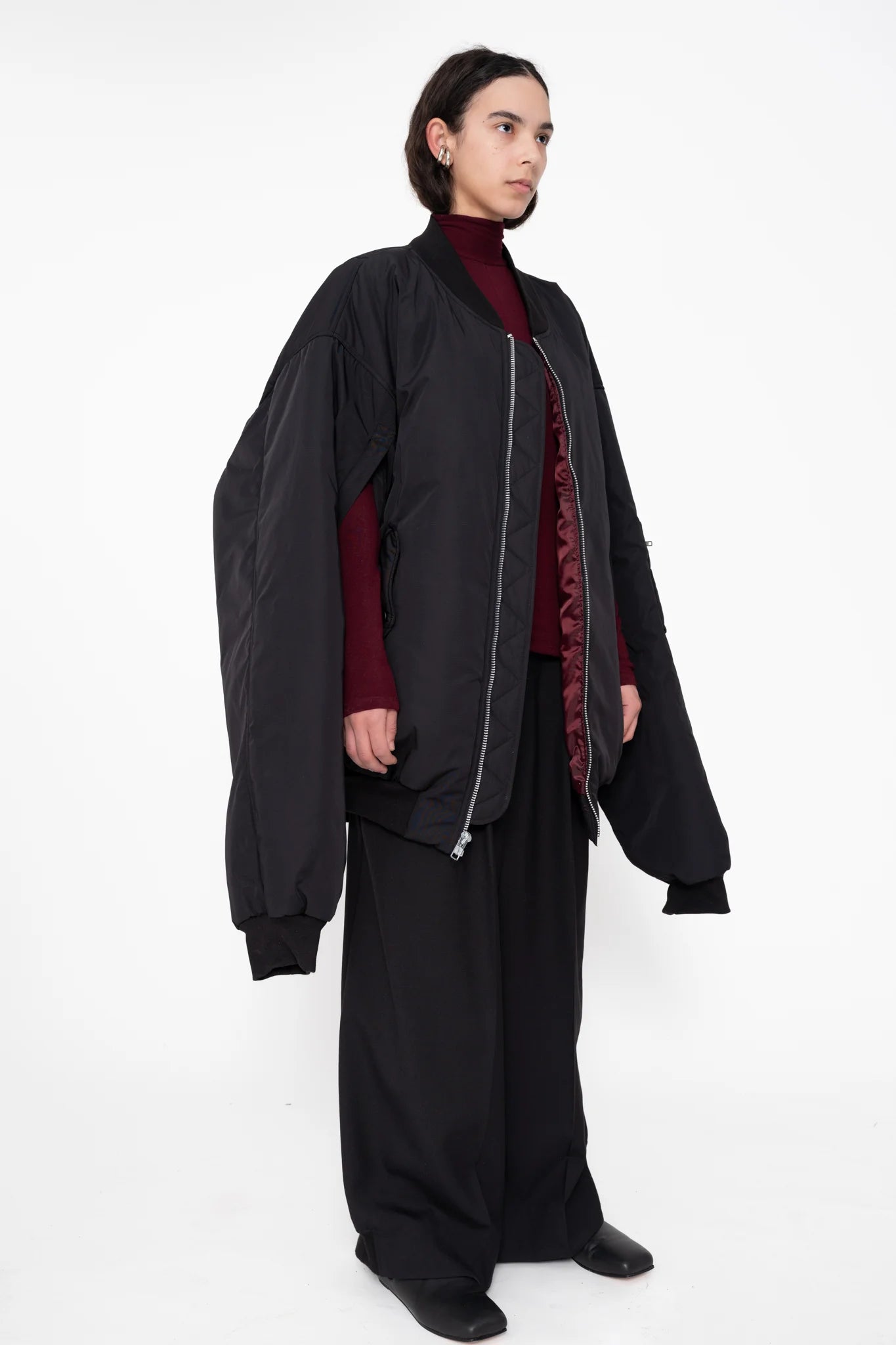 female-wearing-black-oversized-bomber-jacket-with-slits-paired-with-bordeaux-turtleneck-and-black-suit-pants-on-white-background-full-body-three-quarter