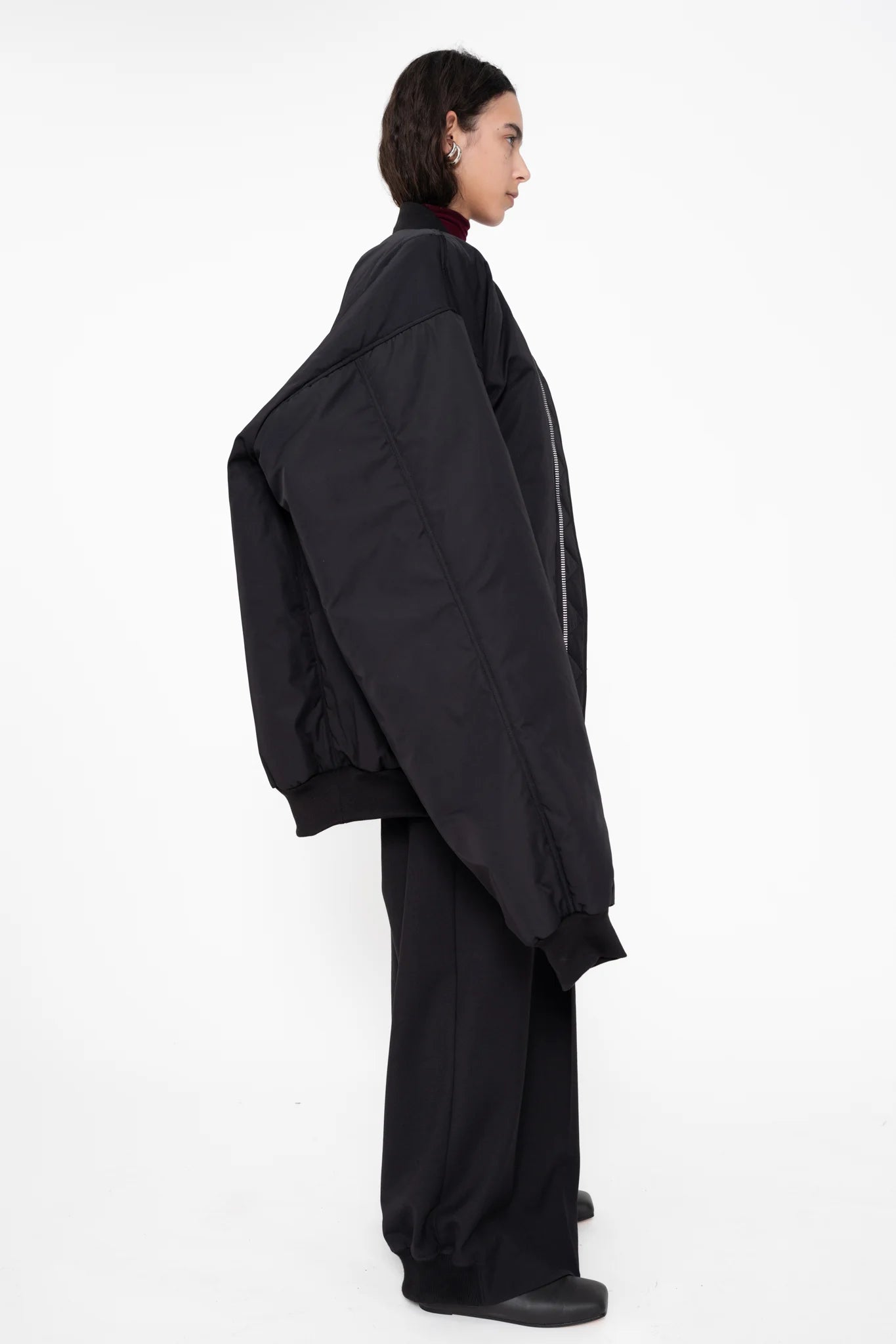 female-wearing-black-oversized-bomber-jacket-with-slits-paired-with-bordeaux-turtleneck-and-black-suit-pants-on-white-background-full-body-side