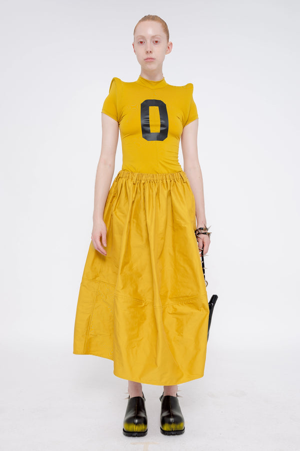 blonde-model-wearing-athletic-tight-mustard-yellow-top-with-foam-shoulders-and-number-zero-mustard-yellow-volume-skirt-holding-glossy-black-banana-tote-spikey-chain-handle-and-black-painted-double-heeled-spikey-mules-has-red-eyes-full-body-front