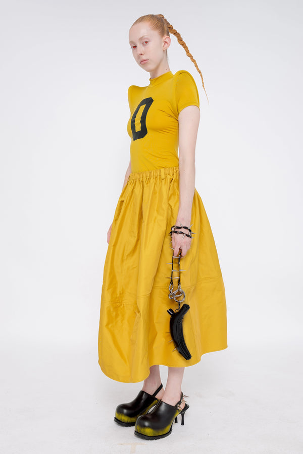 blonde-model-wearing-athletic-tight-mustard-yellow-top-with-foam-shoulders-and-number-zero-mustard-yellow-volume-skirt-holding-glossy-black-banana-tote-spikey-chain-handle-and-black-painted-double-heeled-spikey-mules-full-body-quarter-turn