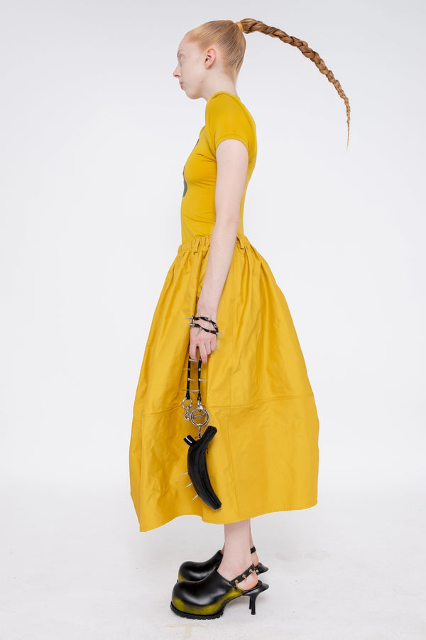 blonde-model-wearing-athletic-tight-mustard-yellow-top-with-foam-shoulders-and-number-zero-mustard-yellow-volume-skirt-holding-glossy-black-banana-tote-spikey-chain-handle-and-black-painted-double-heeled-spikey-mules-full-body-side