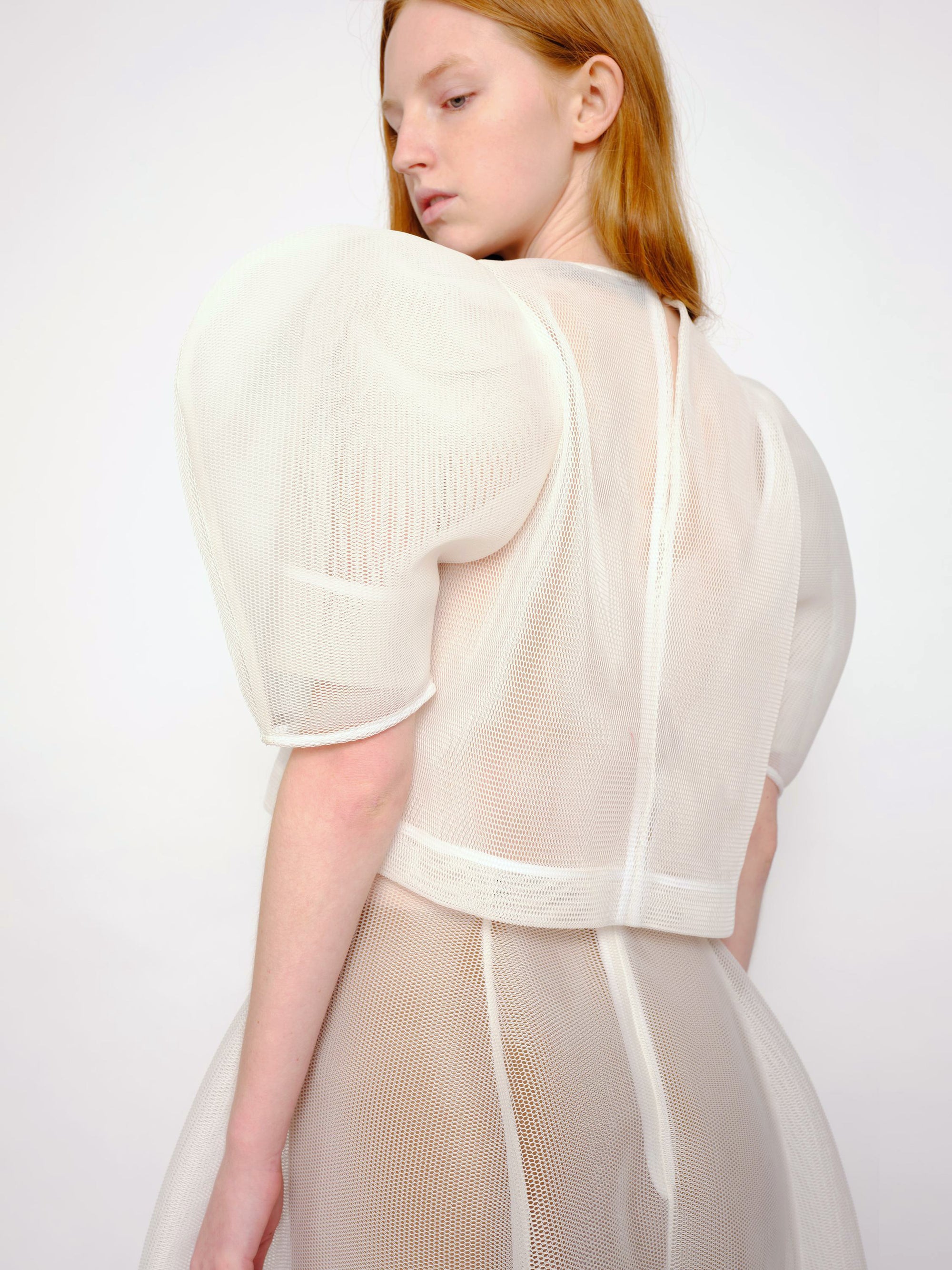 back-angle-of-a-women-with-orange-hair-looking-back-standing-in-front-of-a-white-background-wearing-Melitta-Baumeister-sheer-princess-dress-with-puffy-shoulder-made-in-a-spongy-material