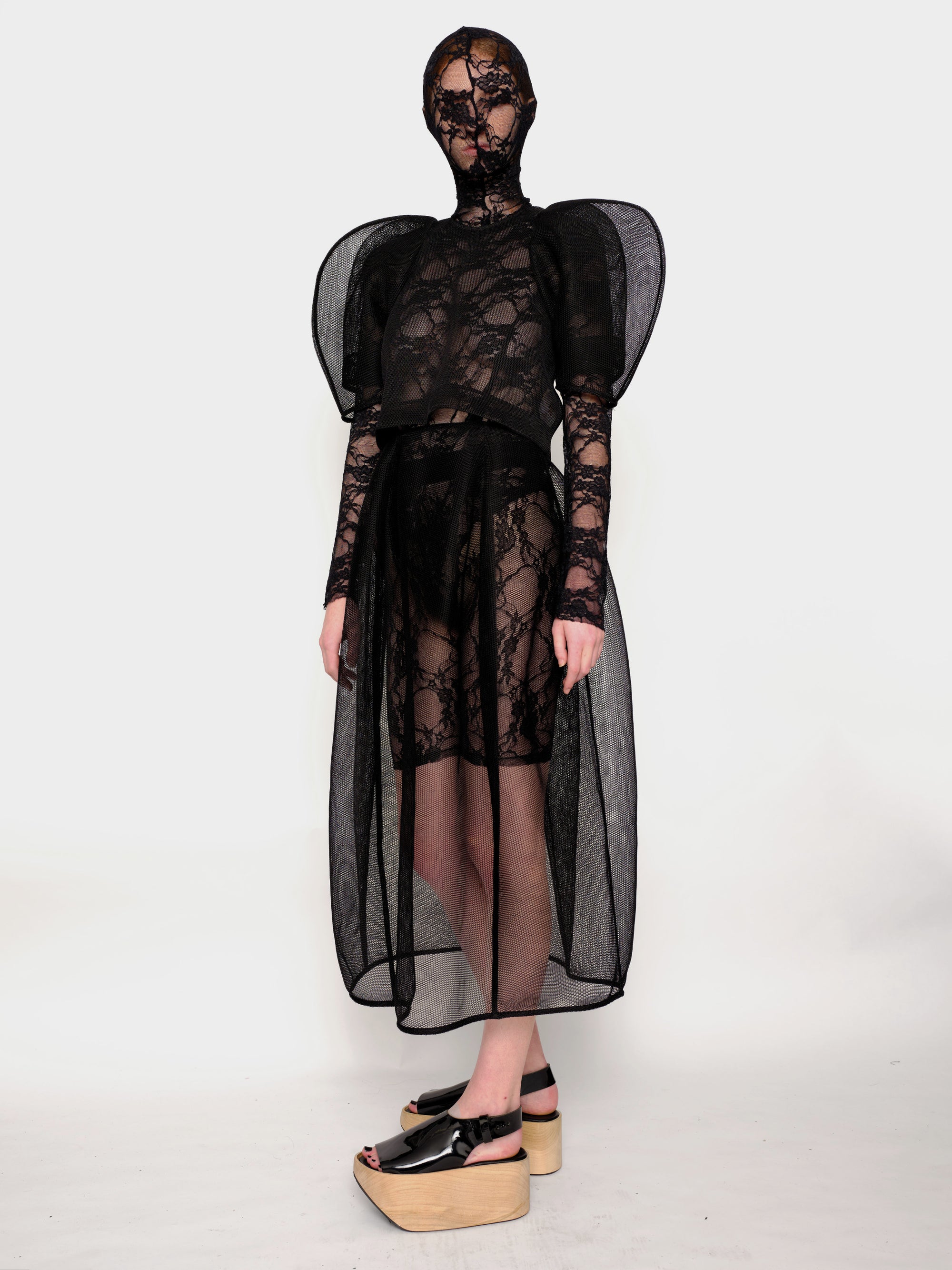 Woman-standing-against-white-background-wearing-Melitta-Baumeister's-black-head-covering-under-layer-covered-by-sheer-lace-dress-with-mesh-fabric-and-puffed-shoulders