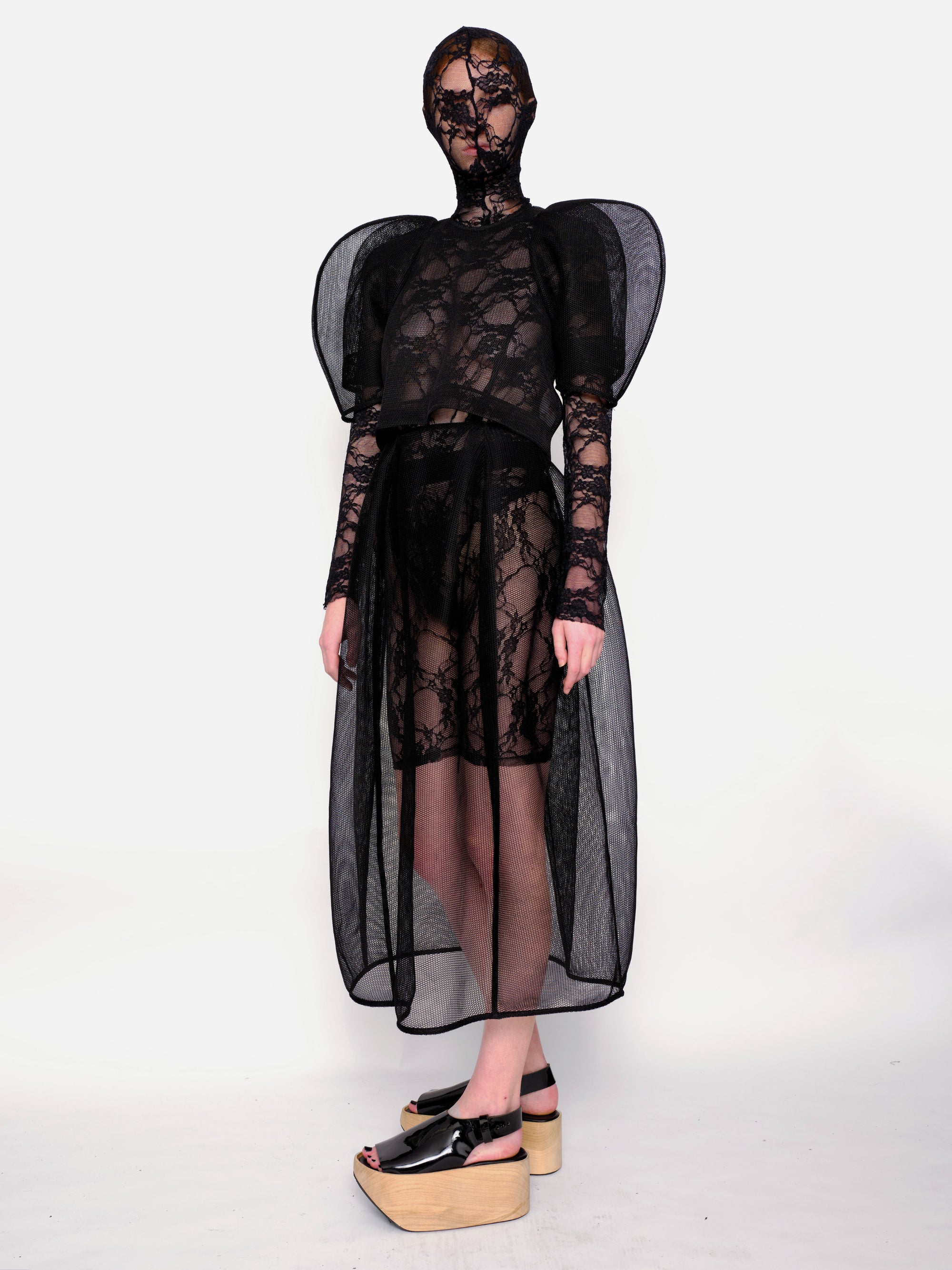 Woman-standing-against-white-background-wearing-Melitta-Baumeister's-black-head-covering-under-layer-covered-by-sheer-lace-dress-with-mesh-fabric-and-puffed-shoulders-and-wearing-platform-sandals