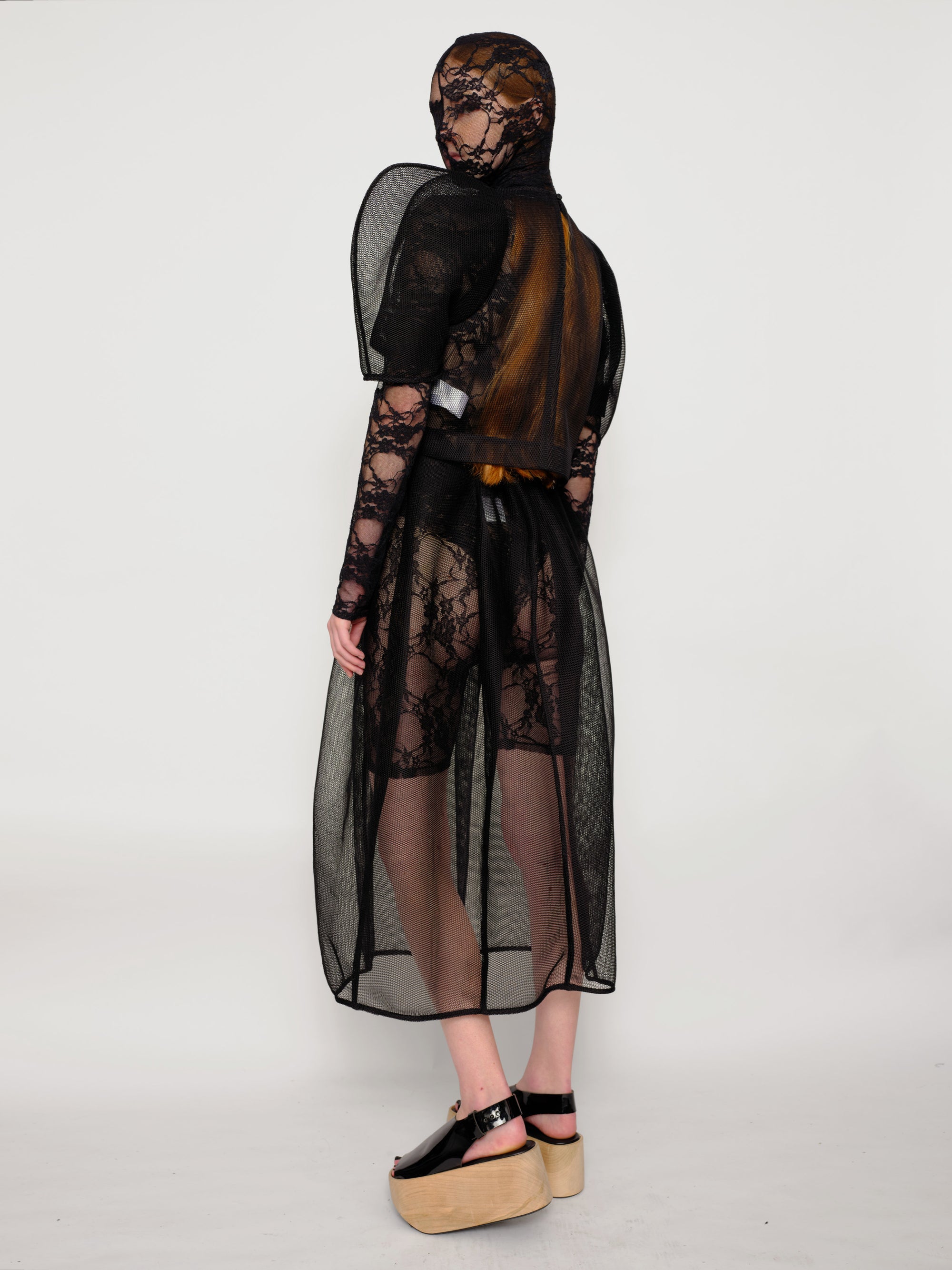 back-angle-of-a-Woman-standing-against-white-background-wearing-Melitta-Baumeister's-black-head-covering-under-layer-covered-by-sheer-lace-dress-with-mesh-fabric-and-puffed-shoulders-and-wearing-platform-sandals